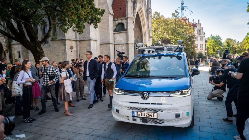 Das Fahrzeug, ein Kleinbus, steht mit der Schnauze zum Betrachter. Rechts und links davon eine Reihe Menschen. Viele davon fotografieren das Fahrzeug. Eher im Hintergrund als Kulisse histroische Gebäude.