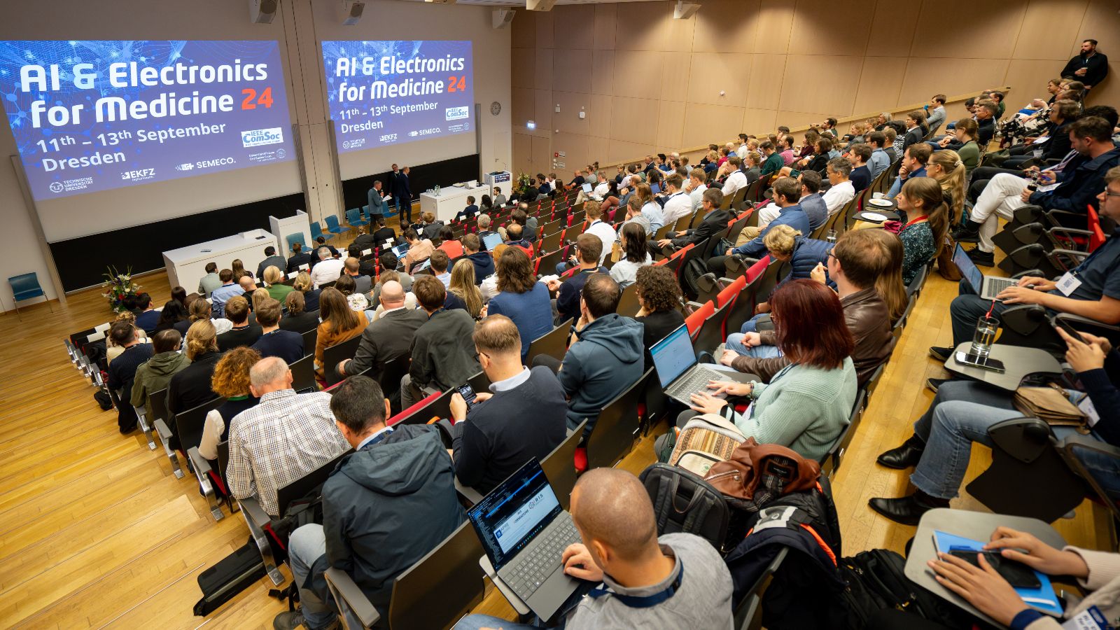 Das Bild zeigt einen Hörsaal voller Menschen. Vorne zwei Leinwände mit identischer Ansicht von Titel und Datum der Veranstaltung