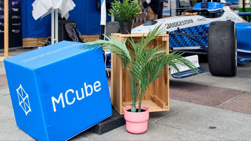ein Teil des MCube Standes auf der IAA 2025: Im Vordergrund ist ein MCube-Würfel, eine Pflanze und eine Holzkiste zu sehen. Im Hintergrund das autonome Rennauto