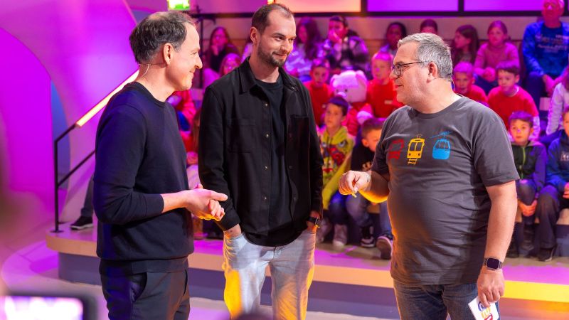 Die beiden MCube-Vertreter beantworten Elton Fragen bei 1, 2 oder 3. Im Hintergrund ist Publikum zu sehen.