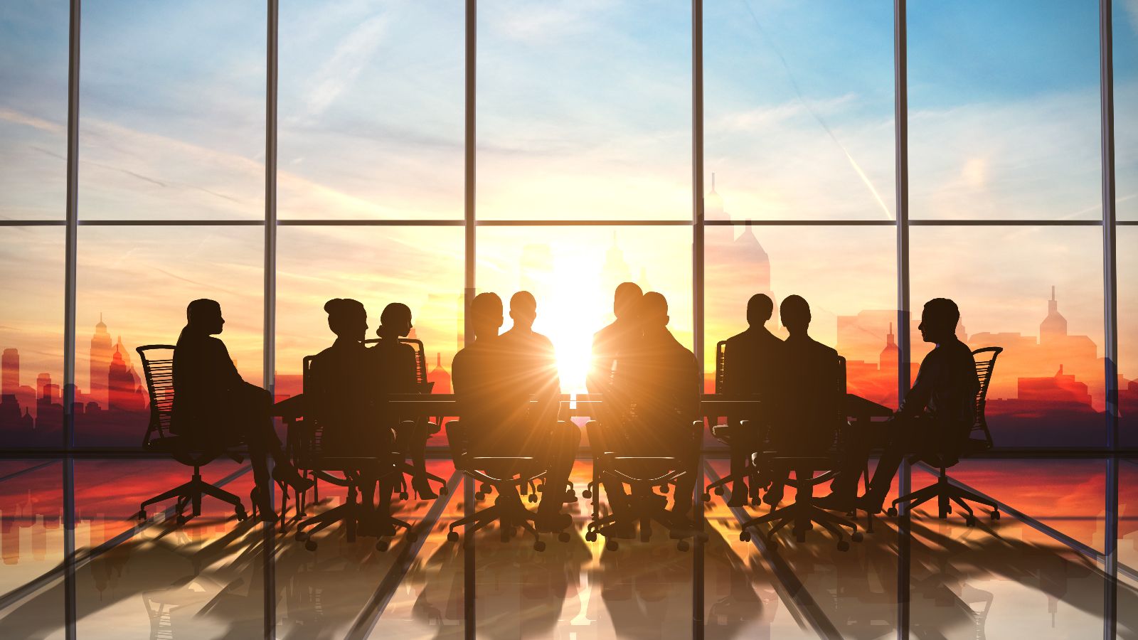 Gruppe sitzt in gläsernem Besprechungsraum. Im Hintergrund geht die Sonne hinter einer Skyline unter.