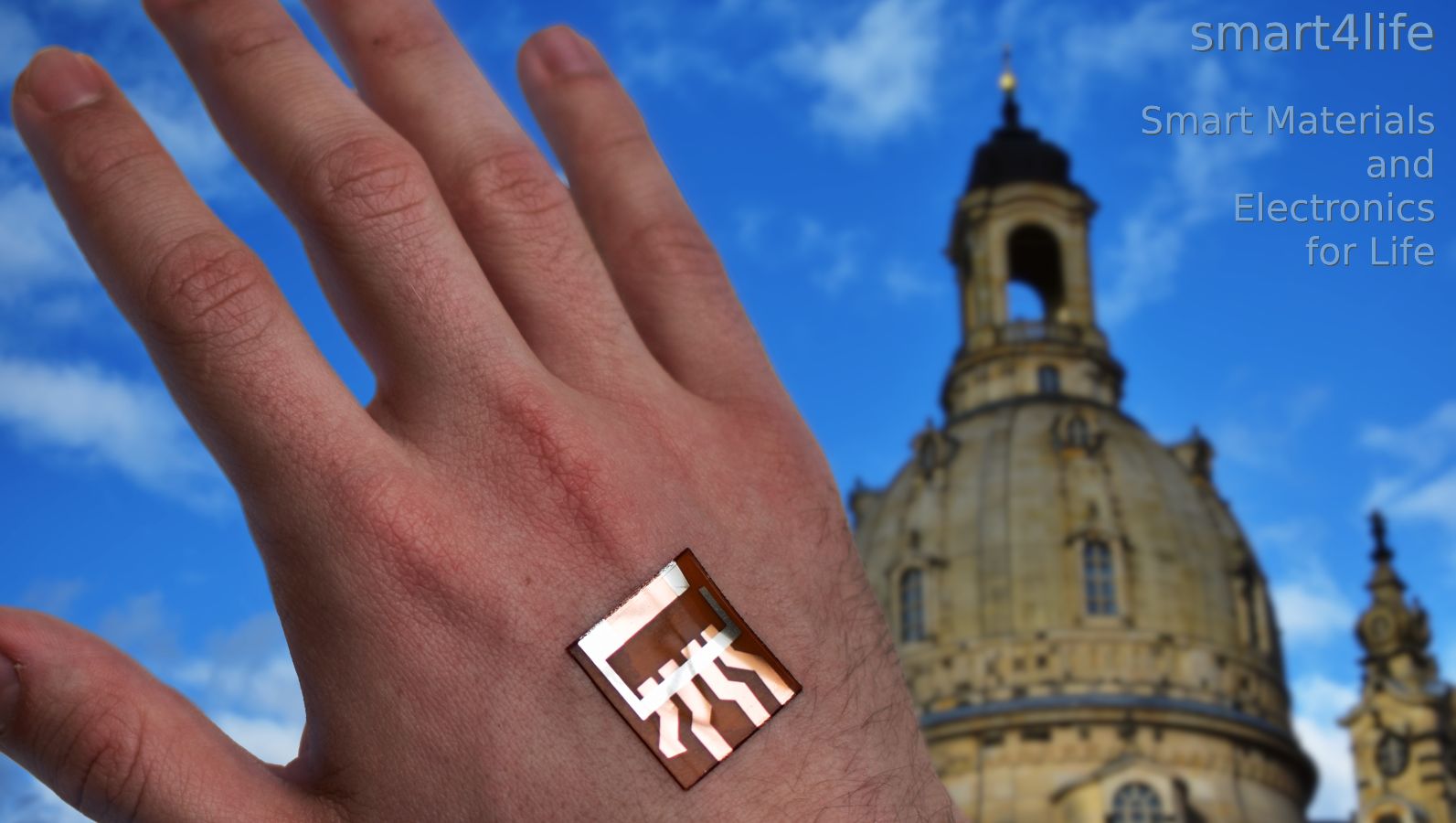 Verbindung elektronischer und biologischer Systeme. Das Zukunftscluster smart4life – Smart Materials and Electronics for Life verbindet elektronische und biologische Systeme. Im Vordergrund ist eine menschliche Hand mit einem flexiblen elektronischen Dünnschichtbauteil zu sehen, im Hintergrund die Frauenkirche als Symbol für die Region Dresden.